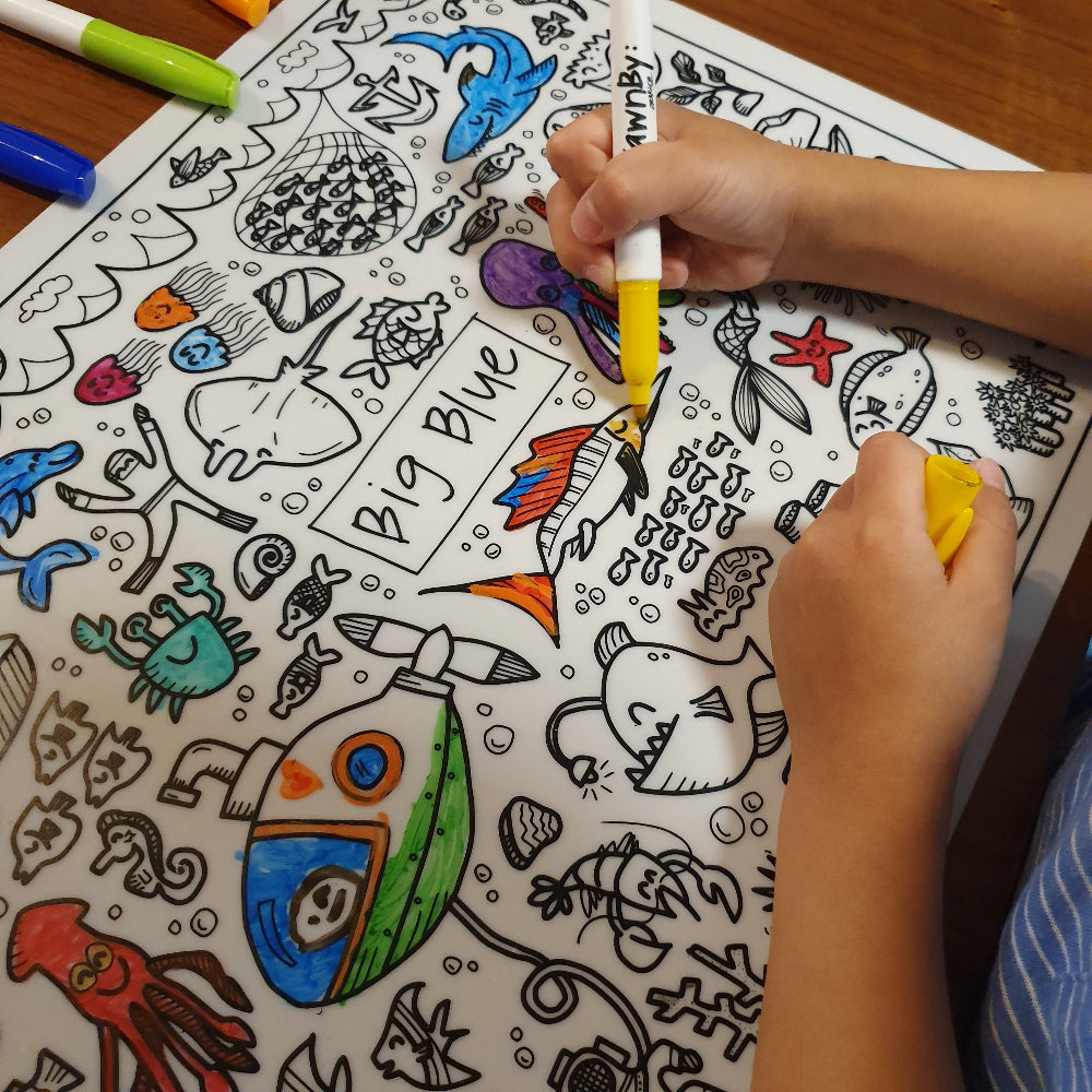 DrawnBy: Big Blue Clouring Mat with underwater-themed illustrations and a child coloring with markers.