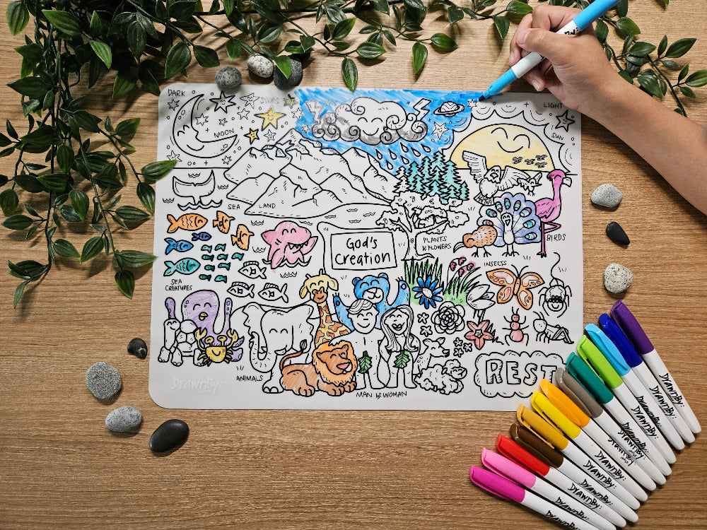 DrawnBy: God's Creation Bible Story Colouring Mat with illustrations of animals and nature, surrounded by markers and stones on a wooden surface.