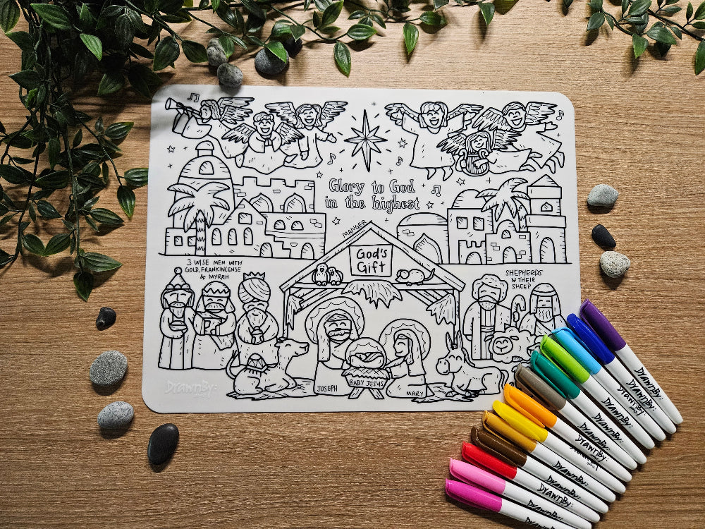 DrawnBy: God's Gift Bible Story Coloring Mat with a nativity scene design on a wooden surface with markers and rocks.
