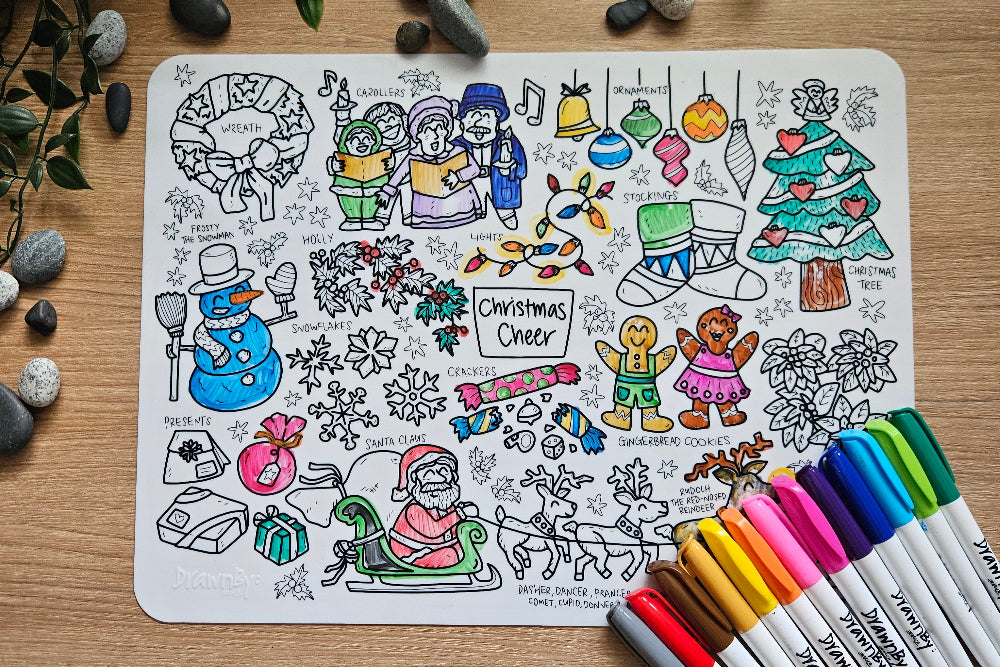 DrawnBy: Christmas Cheer Mat with various holiday designs and markers on a wooden surface.