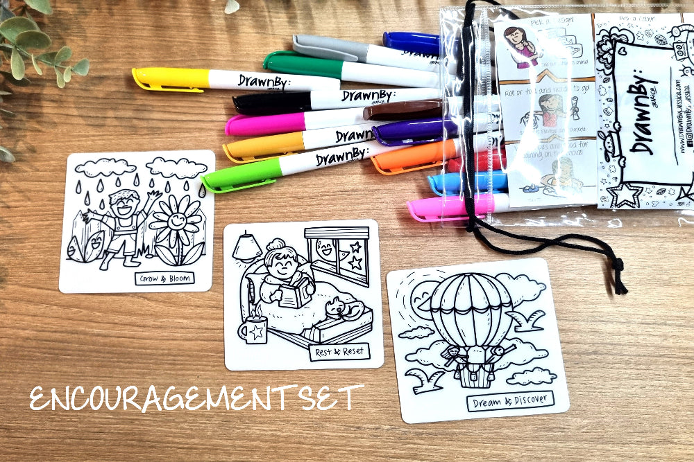 An encouragement art set displayed on a wooden tabletop, featuring three black-and-white sketch cards, a clear zip pouch of colored markers labeled 'DreamBy', and several loose markers in yellow, green, pink, orange, blue, purple, brown, and white. The words 'ENCOURAGEMENT SET' appear in white text at the bottom left.
