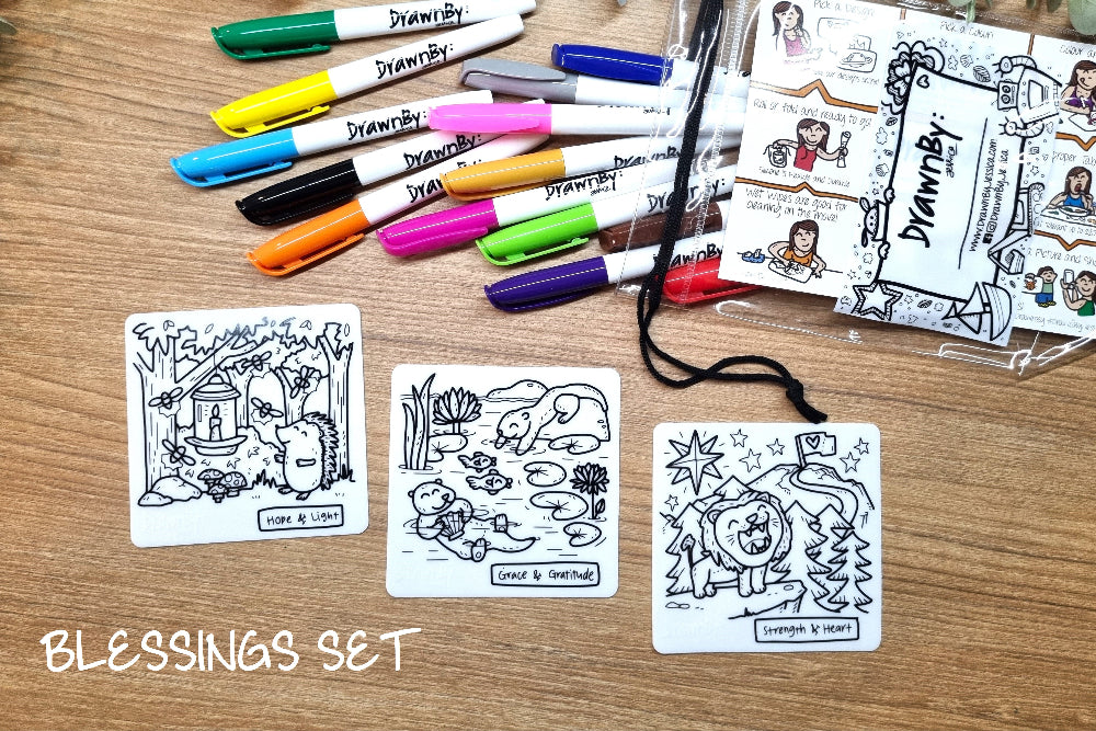 A Blessings Set featuring three small white cards with black line art illustrations, a bundle of colorful drawing markers, and a clear plastic bag of stickers on a wooden tabletop.